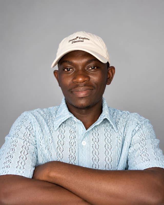 Mojeeb Titilayo in a cream cap and light-blue shirt, arms crossed, smiling against a plain grey background